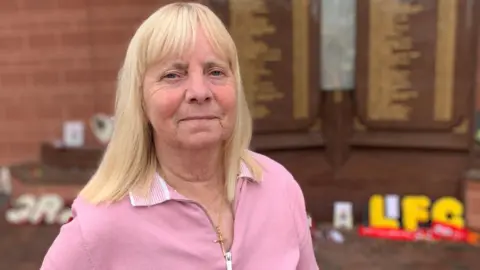 Margaret Aspinall has long blonde hair and is wearing a pink jumper and a gold cross necklace.