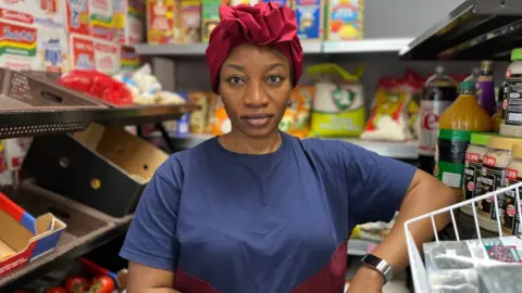 A woman wearing a red hair covering and a blue T-shirt stands in a shop in front of food and drink displayed on shelves. She is looking at the camera but not smiling.