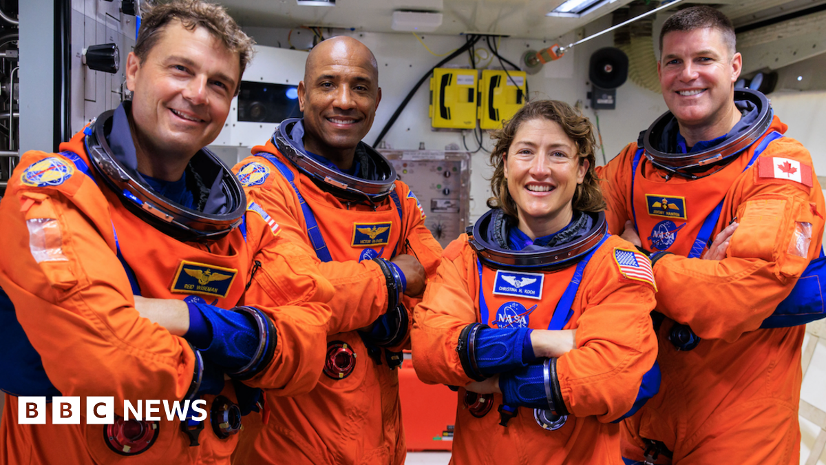 The crew in their orange flight suits standing in what looks like an Orion training room all smiling and with their arms folded