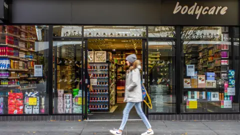 Alamy A person in a grey jacket and blue jeans walks past a shop with 'Bodycare' written on the top-right of the frontage. The store has large glass windows displaying various products such as toiletries, cosmetics, and household items. Shelves of products are visible inside the entrance. Signs on the windows advertise prices and promotions.