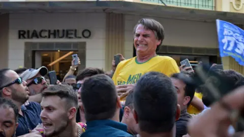 AFP via Getty Images Jair Bolsonaro winces in pain after being stabbed in the stomach during a campaign rally in Juiz de Fora on September 6, 2018. He is wearing a yellow Brazil football shirt. Around him people look distraught