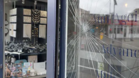 A large cracked glass window at a shop is shown from the exterior, with the reflection of the street outside in it.