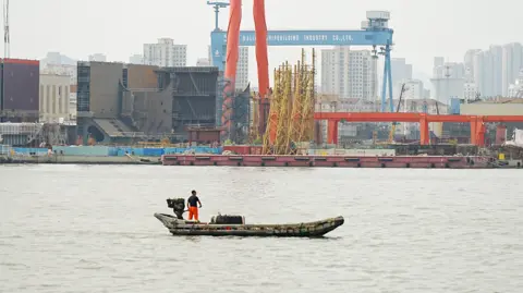 A man stands on a tiny vessel in the Dalian bay - in front of him is the shipyard with huge cranes and other equipment. A tall blue structure has the words Dalian Shipbuilding Industry Co. Ltd on it. 