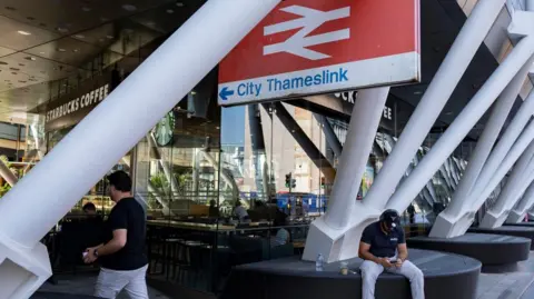 Getty Images A railway station with a large sign bearing the railway symbol and the words City Thameslink. There are white roof support poles in the foreground, and a Starbucks Coffee in the background. A man is sitting below the sign looking at his mobile phone.