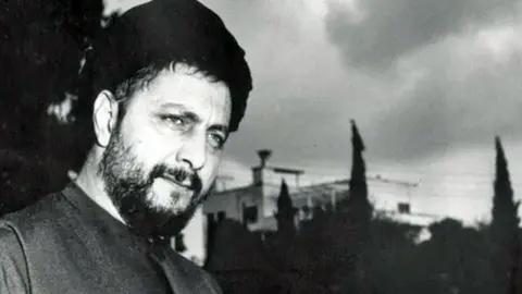 Imam Sadr Foundation A black and white image of Musa al-Sadr. He has a beard, is wearing a hat and is looking thoughtful. There are trees and buildings in the background.