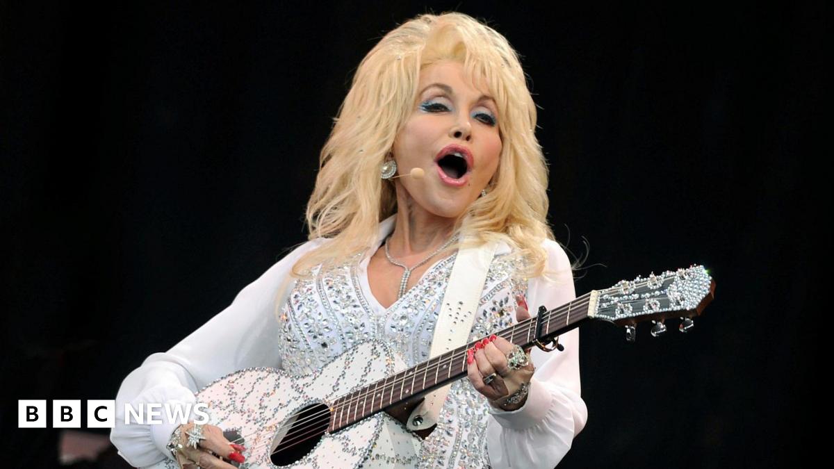 Dolly Parton wearing a white and glittering costume plays a guitar and sings on stage while smiling