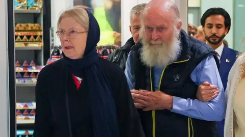 Reuters Older British couple Peter and Barbie Reynolds, who were released from Taliban detention in Afghanistan, walking through arrivals at Heathrow Airport. Barbie is wearing deep blue headscarf and dress, a bearded Peter is wearing a blue shirt and dark blue bodywarmer.