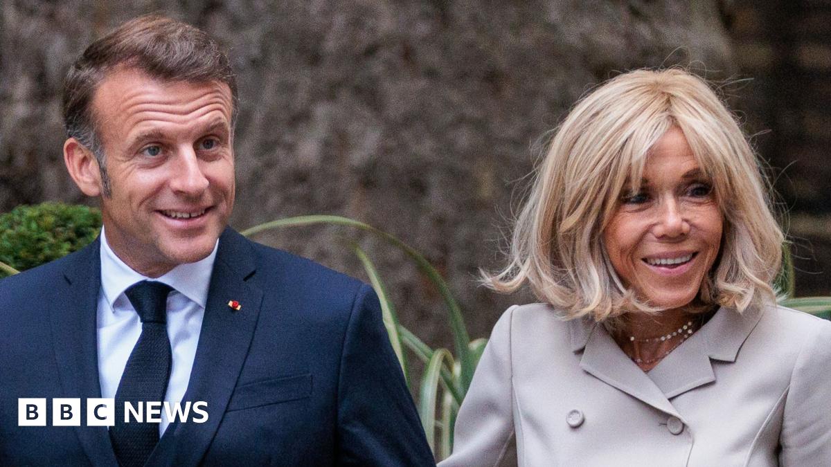 French President Emmanuel Macron and First Lady Brigitte Macron arriving in Downing Street in July. The image depicts the couple walking arm in arm. Mr Macron is dressed in a dark blue suit, white shirt, and dark tie. Mrs Macron is wearing a light-colored, double-breasted jacket suit. They are surrounded by greenery, including bushes and plants with long leaves