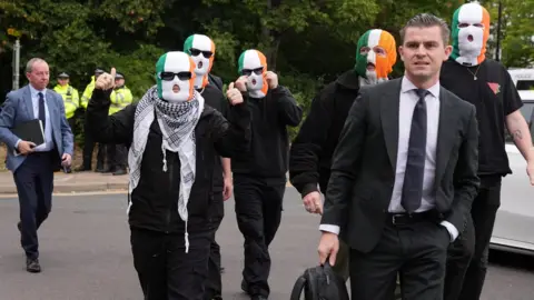 Reuters A group of people arriving to Woolwich Crown Court. Five of the men are wearing green white and orange tricolour balaclavas. They are all dressed in black.