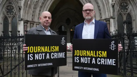 PA Media Barry McCaffrey and Trevor Birney outside the Royal Courts of Justice, London, in October. They are holding black signs with yellow and white writing that say 'Journalism is not a crime'. There are black iron gates behind them.
