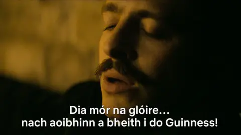 Netflix A still from the show, that shows Anthony Boyle as Arthur Guinness with Irish subtitles "Dia mór na glóire ... nach aoibhinn a bheith i do Guinness!"