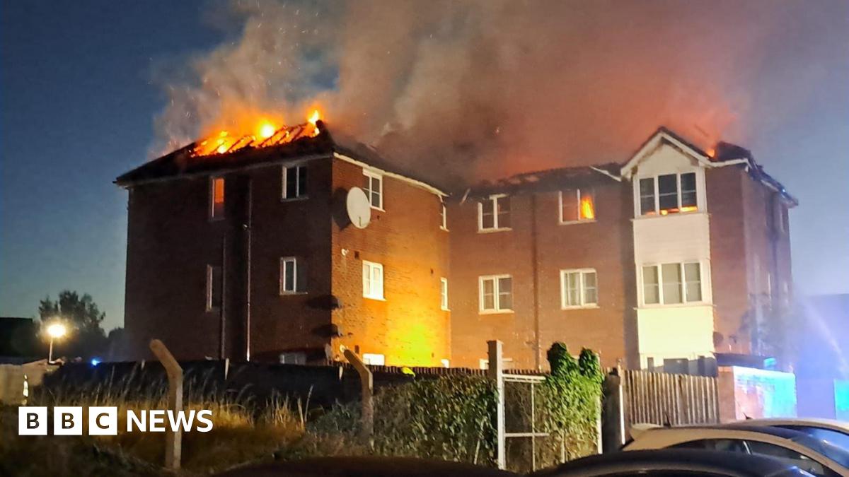 A  fire engulfs the roof of a three-storey red-brick block of flats at night. Flames and thick smoke rise into the sky, with several windows glowing orange from the blaze. The lower floors remain intact, and parked cars are visible in the foreground.