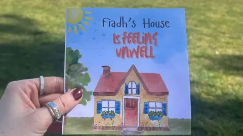 Síofra Harkin A book with a picture of a house on it with the title: 'Fiadh's House is feeling unwell' accompanied by a picture of a yellow and red house with a number of cracks visible on its front. 