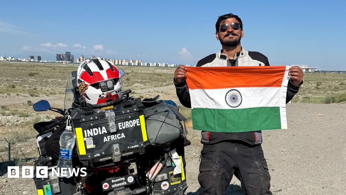 A photograph of a man standing next to a bike while holding the flag of India