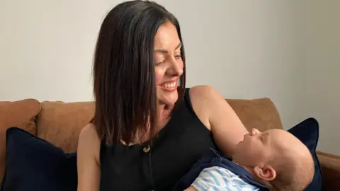 Vanessa Clarke/BBC Mum Rachel smiles down at her baby son Gabriel, sat on her sofa at home. She has dark, shoulder-length hair and is wearing a black top.