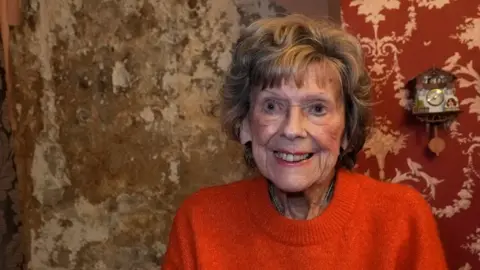 Margaret Chappell a 93-year-old woman whose bright smile and immaculate appearance with neatly permed light brown hair and tasteful make-up contrasts with the wall behind her which is consumed by brown mould with plaster peeling off it. 