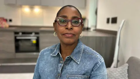 BBC Patricia Ogunfeibo, wearing shaped dark red coloured glasses, hooped earrings and a denim collared shirt, is sitting with a kitchen worktop in the background behind her