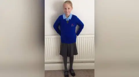 Rachel Cowley A young girl with blonde hair wearing a blue school uniform, a school skirt and black school shoes. She is stood in front of a radiator leaning on a white wall.