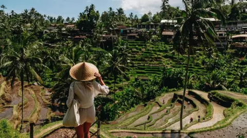 Getty Images Back view of a woman in white shirt and shorts wearing an Asian conical hat. She is holding the rim of her hat and looking at vast terraced rice fields and small houses in the distance.