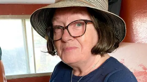 Sheila O'Donnell A woman wearing a straw sun hat and glasses smiles at the camera. She has brown hair in a bob, and is wearing a navy blue top and a silver necklace. In the background is a wall painted brick red and a window.