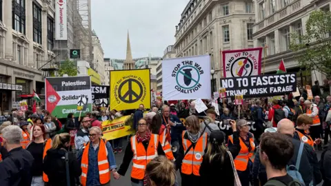 BBC People wearing orange reflective jackets holding hands at the front of the march. Behind them are crowds of people some holding banners, including some that read: Palestine Solidarity Campaign, Campaign for Nuclear Disarmament, Stop the War Coalition, No to Fascists, and Stand Up to Racism. 