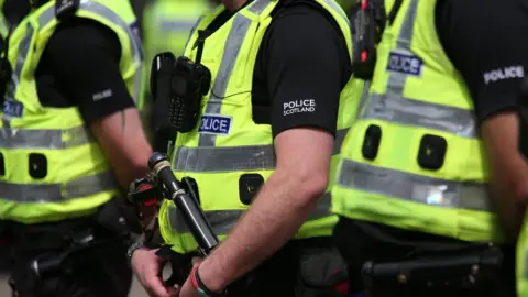 PA Media Several Police Scotland officers in uniform. They are wearing high-visibility yellow vests, black polo shirts with the "Police Scotland" logo, and various equipment including radios and a baton. Other officers are visible in the background.