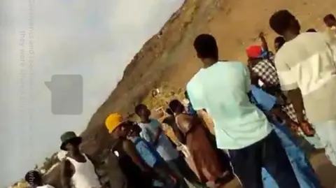 James Nunan/Facebook A still taken from James Nunan's Facebook Live broadcast. It shows people, believed to be migrants, gathered on a beach.