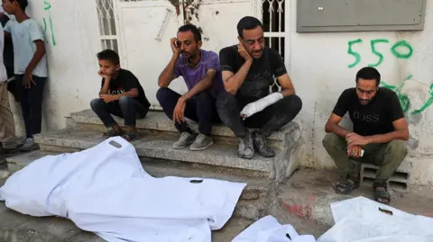Reuters Mourners sit next to the bodies of Palestinians killed in overnight Israeli strikes, outside al-Shifa Hospital in Gaza City (2 September 2025)