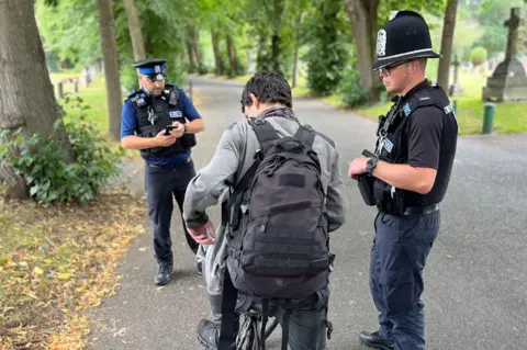 Two police officers in uniform stand in a park surrounded by trees next to a man on a bike, wearing grey with a black rucksack.
