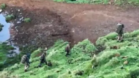 New Zealand Herald A blurry screenshot of a video taken on a phone showing four people pictured from above walking in the wilderness carrying rucksacks and wearing outdoor gear, in October 2024.