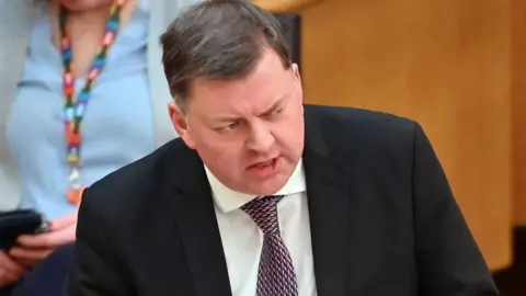 Getty Images Colin Smyth, a man with dark hair, speaking in the Scottish Parliament. He is wearing a dark suit, white shirt and purple tie. There is wooden panelling and a person using a phone in the background. 