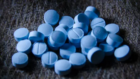 Getty Images Several round bright blue pills scattered on a dark grey and blue fabric