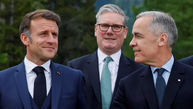 A suited Emmanuel Macron smiles on, as Mark Carney - also in a suit - looks over at him. Keir Starmer, wearing glasses and a suit, stands behind them both while smiling