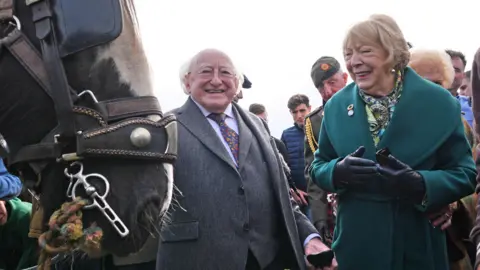 PA Media A brown horse is in the foreground. Michael D Higgins and his wife Sabina stand beside it. He is wearing a grey suit and a patterned tie. She is wearing a dark green coat and leather gloves.
