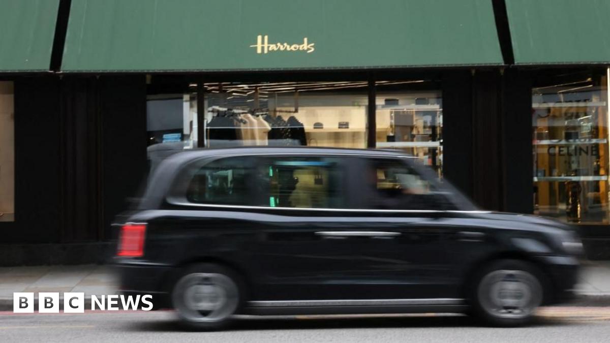 A blurry black cab rushes past the Harrods shop front, with green awnings with "Harrods" printed in gold on one.