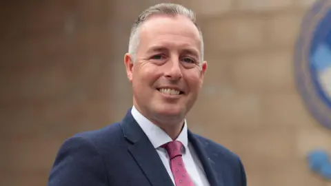 PA Media Paul Givan ;looking at the camera and smiling. He has a navy blazer and pink tie. 