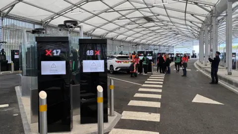 Simon Jones/BBC Two finger printing machines in the foreground of a hanger at Eurotunnel in Folkestone with cars and people in the background.