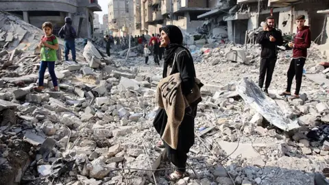 AFP via Getty Images A distraught-looking woman wearing black stands amid rubble and wreckage. There are crowds in the distance moving down a road. A young girl looks on, also looking sombre. 