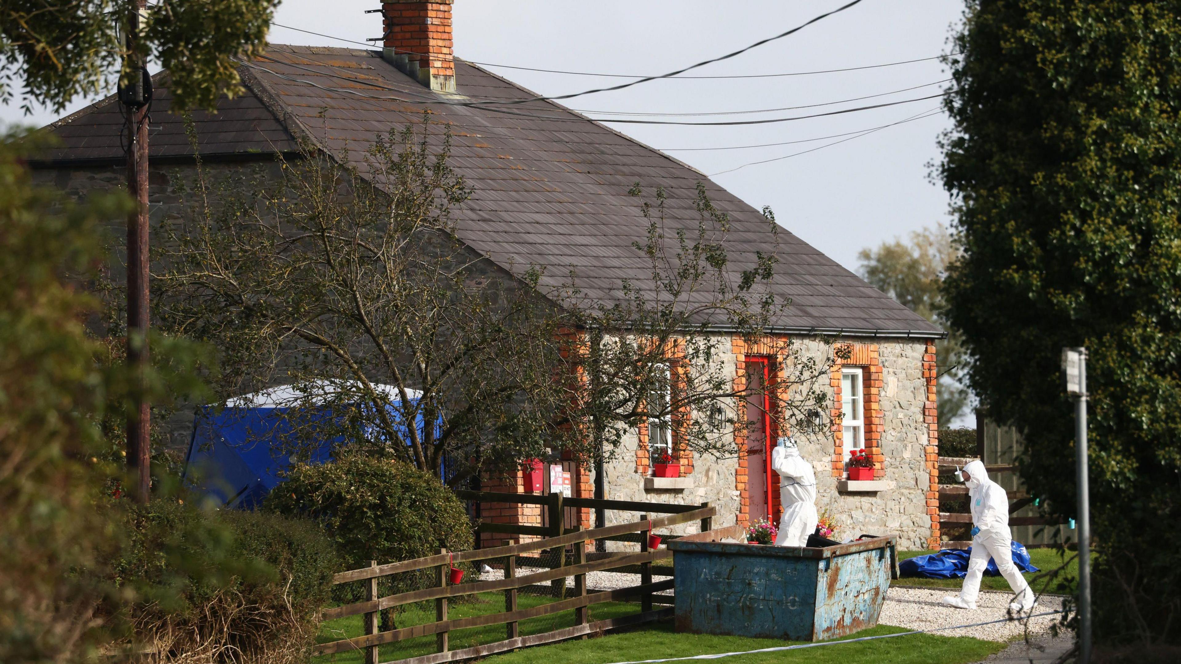 Forensic investigators in white hazmat suits entering a detached stone cottage on a rural road in County Louth.  A blue tent has been erected at the side of the house and there is a blue skip at the front of the property.  The red-framed front door is open.  There are red flower boxs on the front windowsills. 