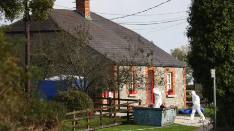 Peter Morrison/PA Wire Forensic investigators in white hazmat suits entering a detached stone cottage on a rural road in County Louth.  A blue tent has been erected at the side of the house and there is a blue skip at the front of the property.  The red-framed front door is open.  There are red flower boxs on the front windowsills. 
