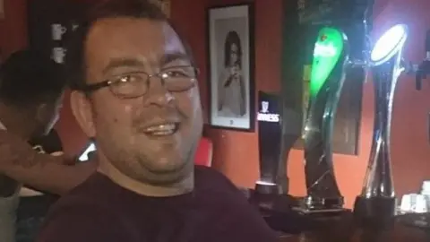 Family handout Head and shoulders of Danny Aitchison sat in a bar next to three pumps. He is smiling and has short dark hair and wire framed glasses and a burgundy tshirt