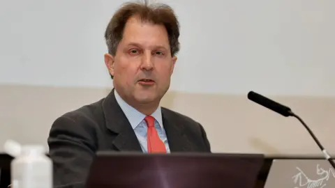 PA Media Angus McCullough KC has dark short hair and is sitting behind a microphone.
He is wearing a grey suit, pale blue shirt and red tie.
