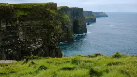 Getty Images A vista of the Cliffs of Moher. The photo is taken from an edge of grass leading to a drop. The sky is cloudy. 