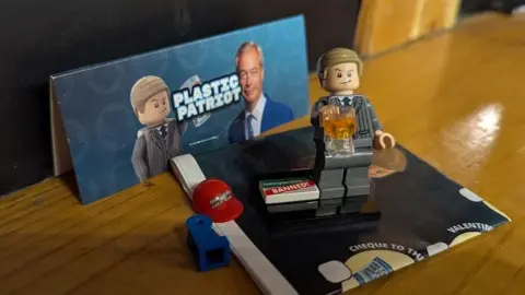 Nigel Farage Lego figure clutching a beer, against a backdrop of "plastic patriot" packaging