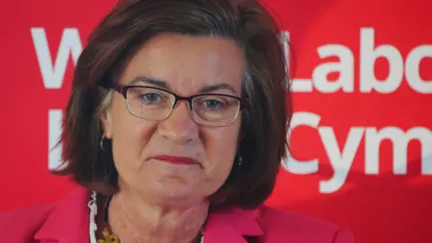 PA Media Eluned Morgan speaking in front of a red Welsh Labour banner, wearing a pink jacket. She has a gold chunky necklace on, purple framed glasses and dark brown shoulder-length hair. 