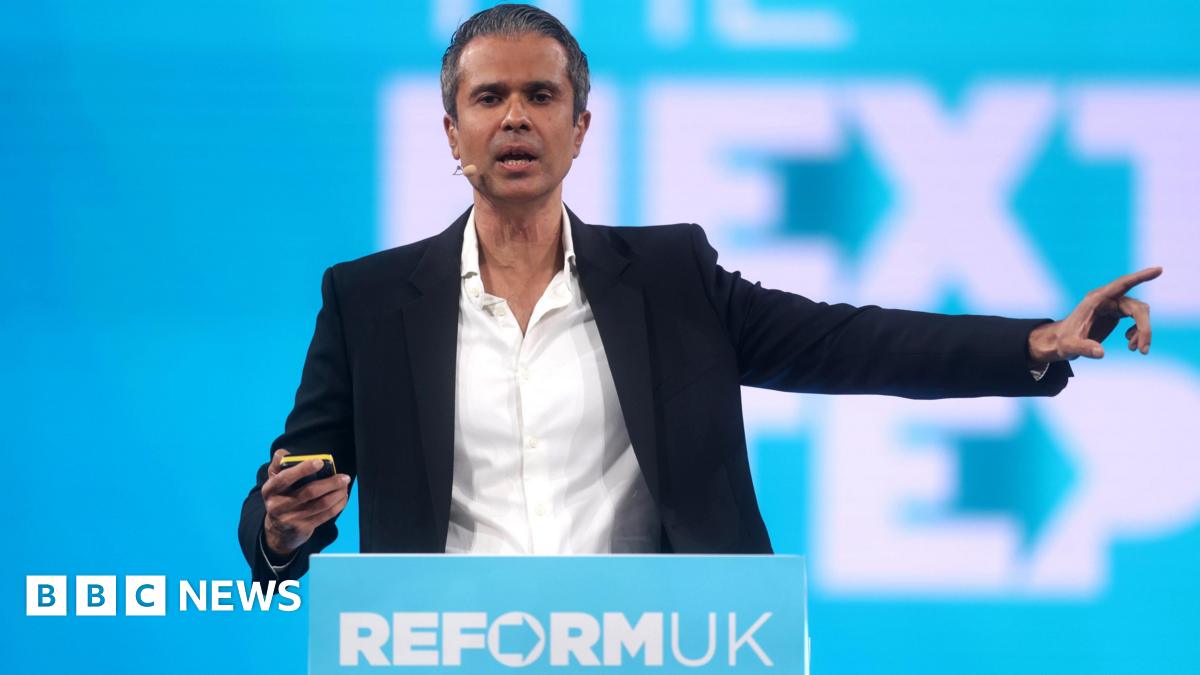 Aseem Malholtra, in a white shirt and dark suit, stands behind a Reform UK podium