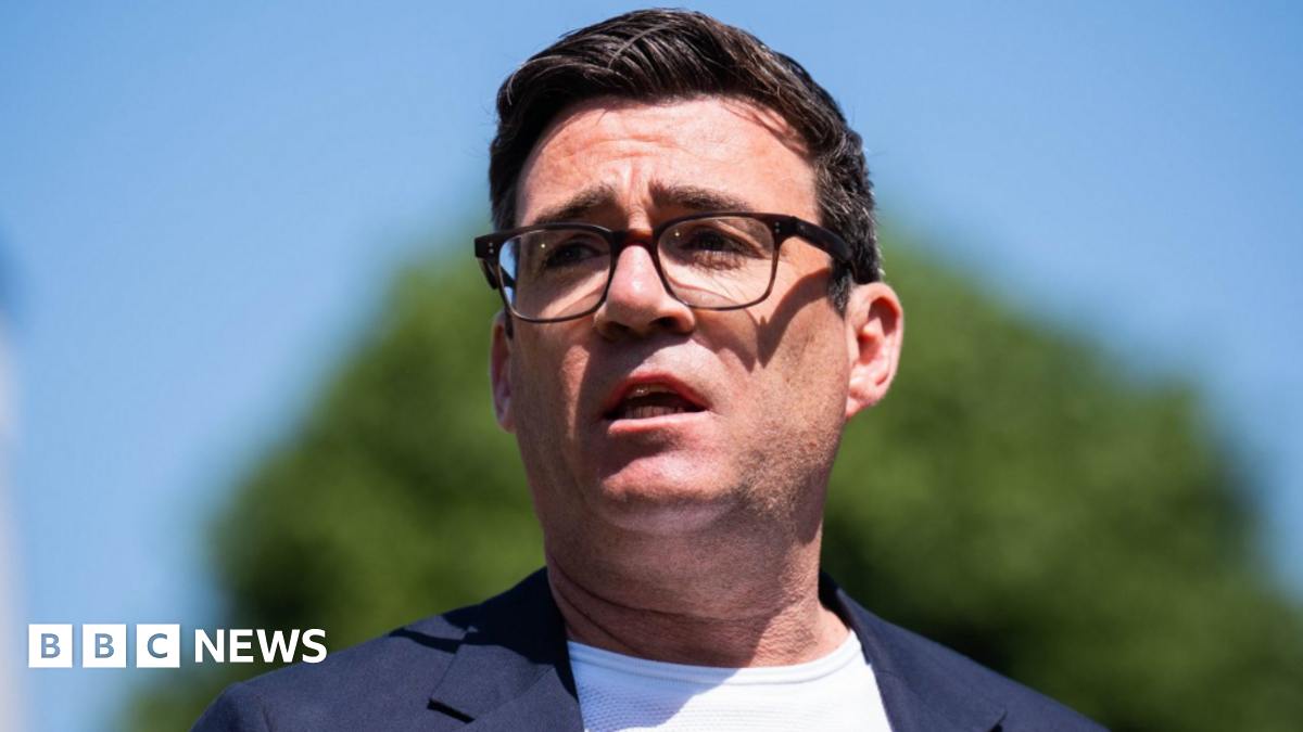 Headshot of Andy Burnham, Mayor of Greater Manchester, outside. He has a tree behind him and is wearing a navy suit and a white t-shirt, His dark hair is styled into a small quiff and he wears dark rimmed spectacles.