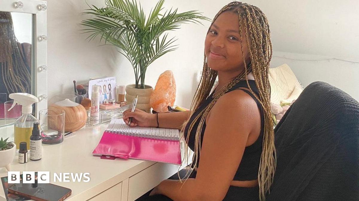 A woman with long braided hair sits at a desk and vanity table in her bedroom, writing notes in her university study book