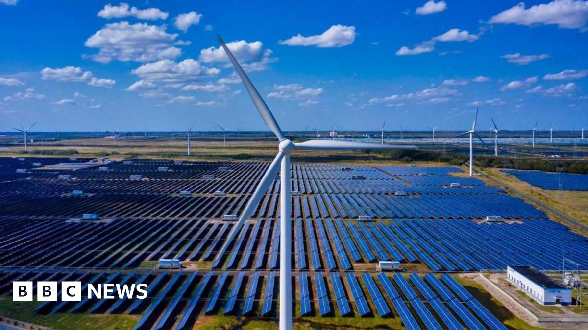 A close-up shot of a wind turbine, behind which are rows and rows of solar panels, with more wind turbines in the distance. The sky is blue with a few clouds.