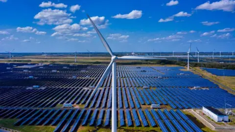 European Photopress Agency A close-up shot of a wind turbine, behind which are rows and rows of solar panels, with more wind turbines in the distance. The sky is blue with a few clouds.
